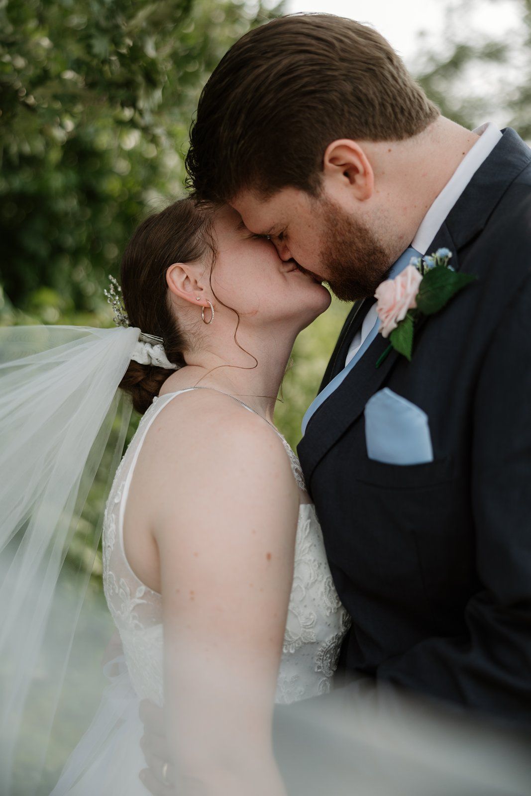 A romantic wedding moment captured outdoors as newlyweds share a tender kiss, with the bride's veil flowing in soft focus.