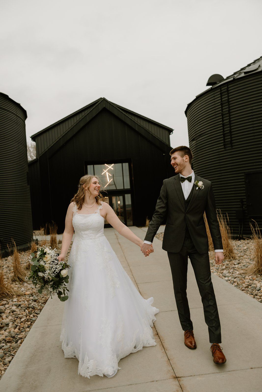A wedding couple walks hand in hand along a path beside modern black barn buildings on their special day.