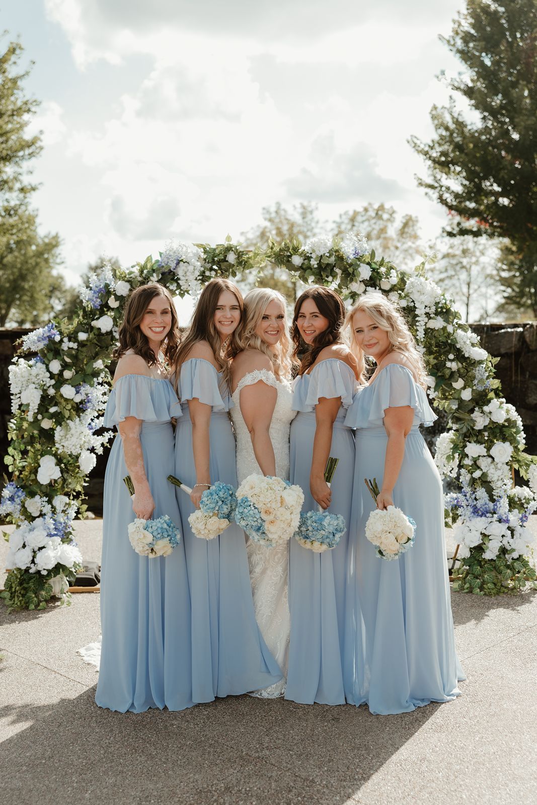 Bridesmaids in flowing powder blue dresses pose with bride under a floral arch.