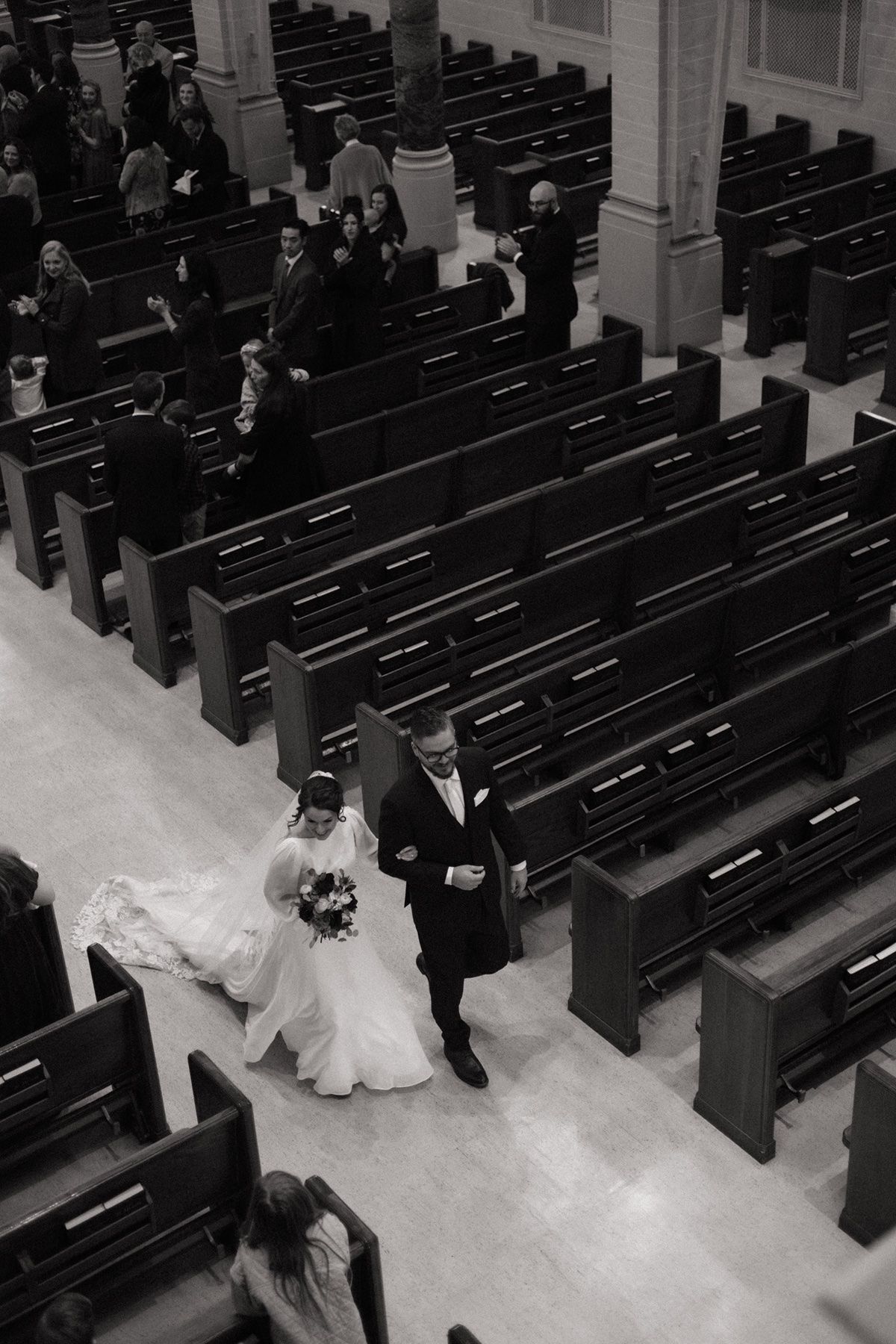 Black and white aerial view of bride and groom walking down church aisle during wedding ceremony with wooden pews.