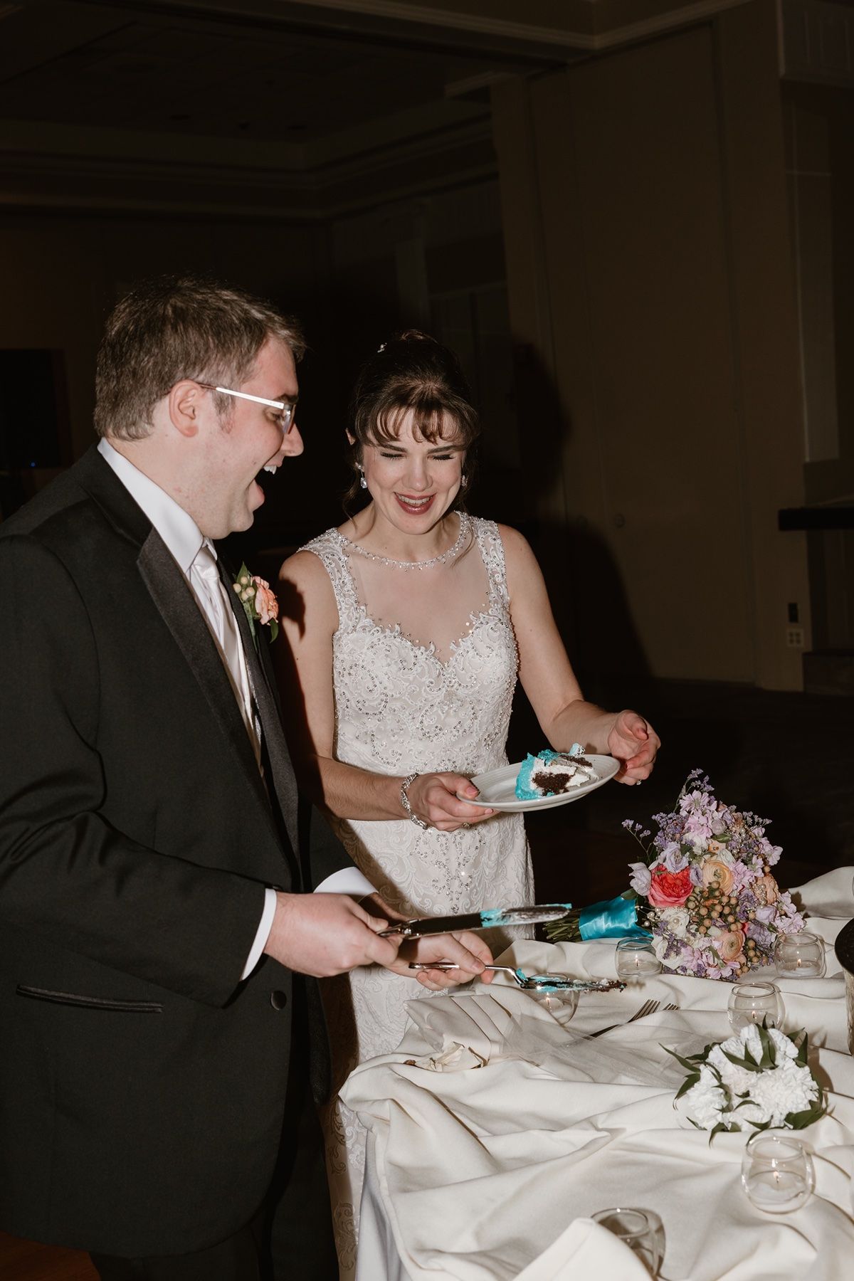 Bride and groom smiling while cutting wedding cake at reception table with floral bouquet nearby.