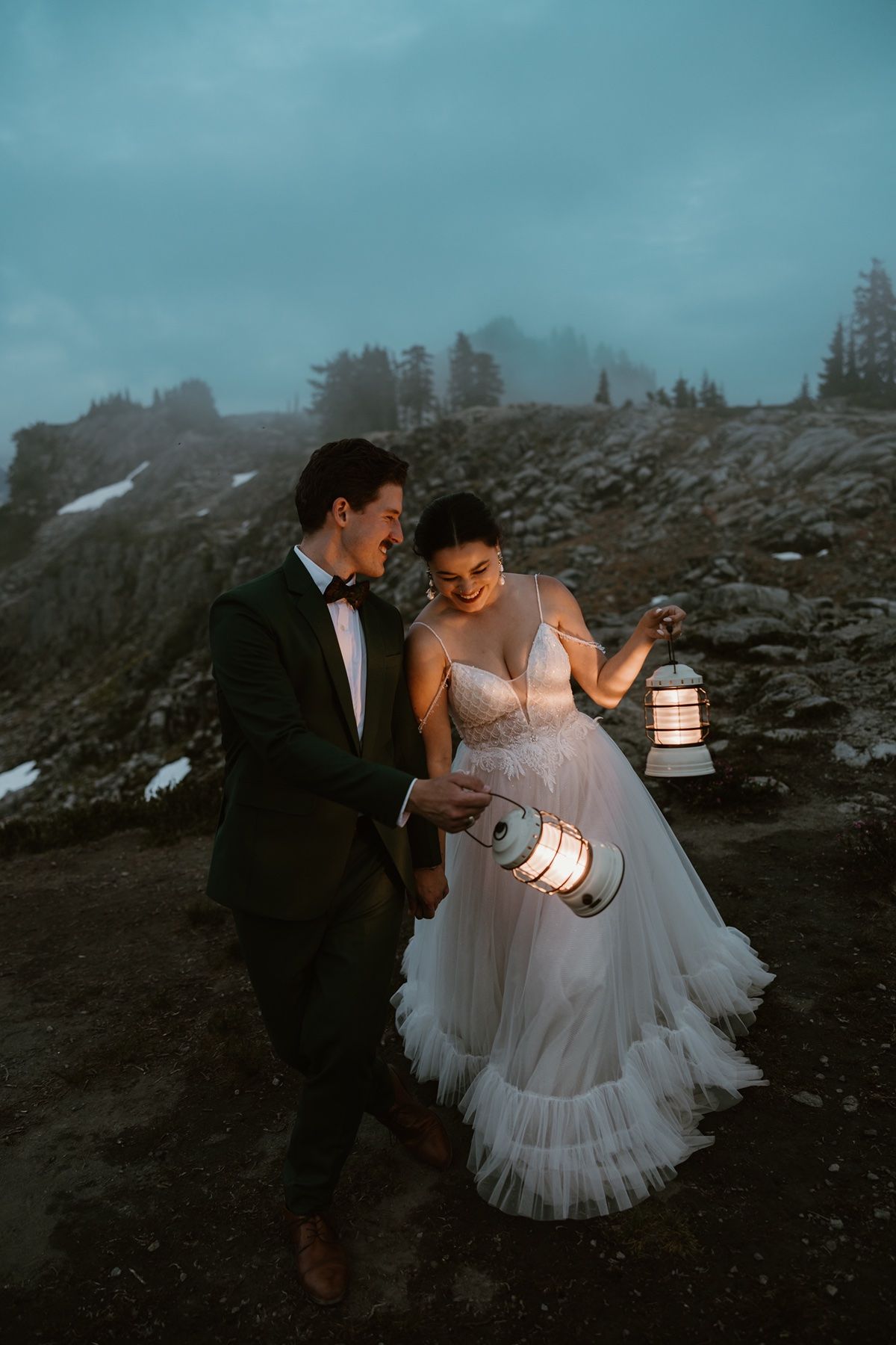 Bride and groom walk together at dusk holding glowing lanterns in a misty mountain landscape during their adventure elopement