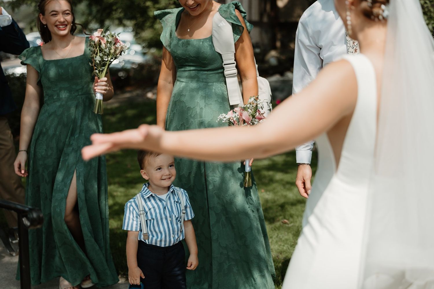 A young boy in striped shirt stands with attendants in emerald green dresses at an outdoor wedding ceremony.