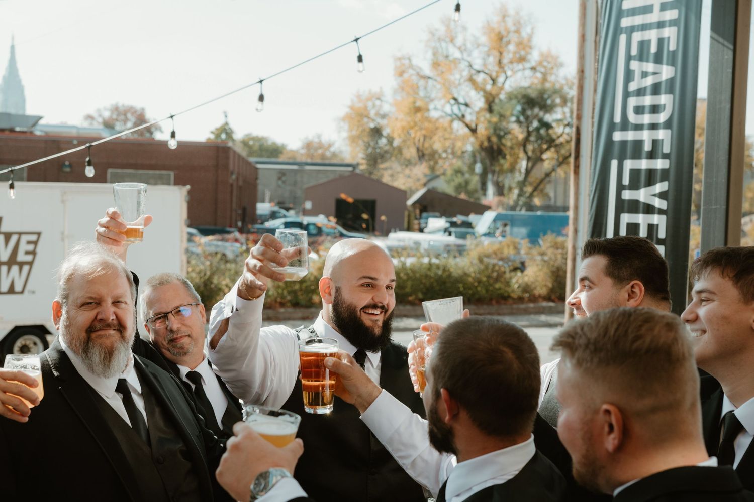A group of well-dressed guests raise beer glasses in a toast at an outdoor celebration.