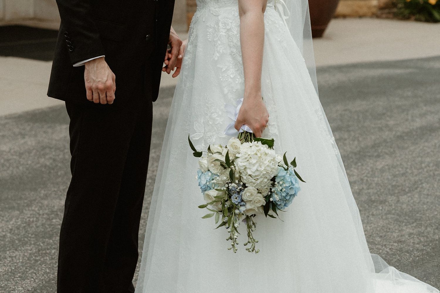 A wedding couple stands together outdoors while holding a white and light blue floral bouquet.