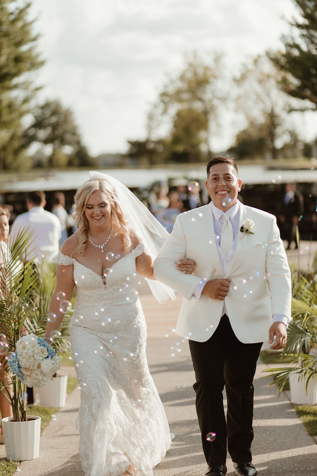 A joyful wedding couple walking together outdoors, with the bride in white lace and groom in white jacket.