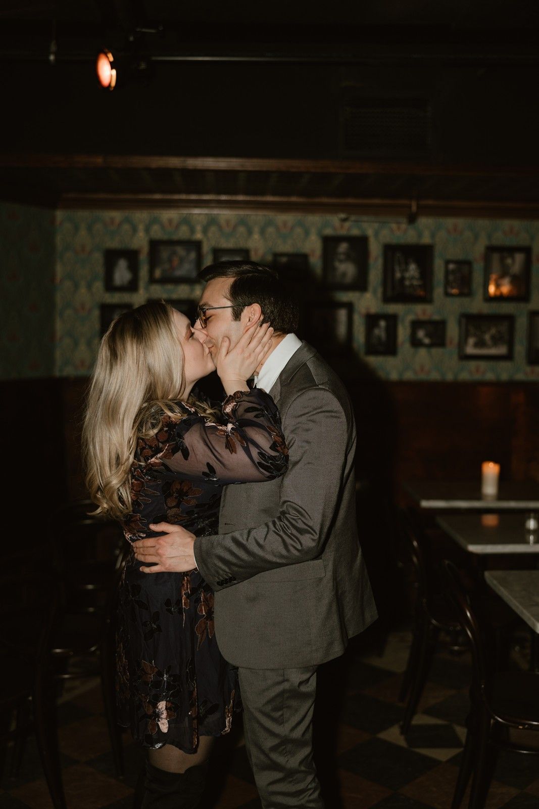 A romantic evening embrace in a dimly lit vintage bar with framed photographs on the wall.