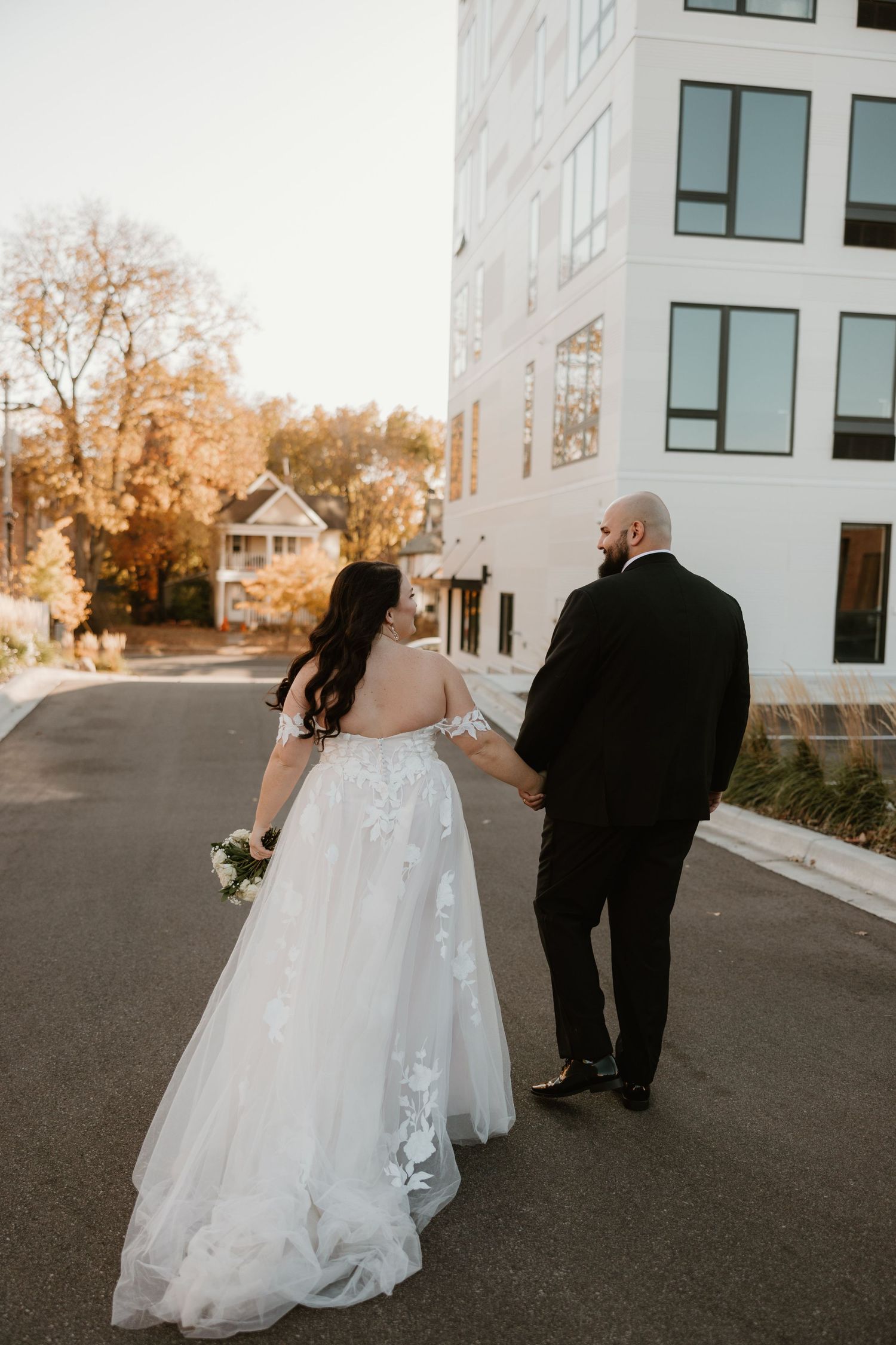 A couple in wedding attire walks down a street at sunset beside a modern white building with large windows.