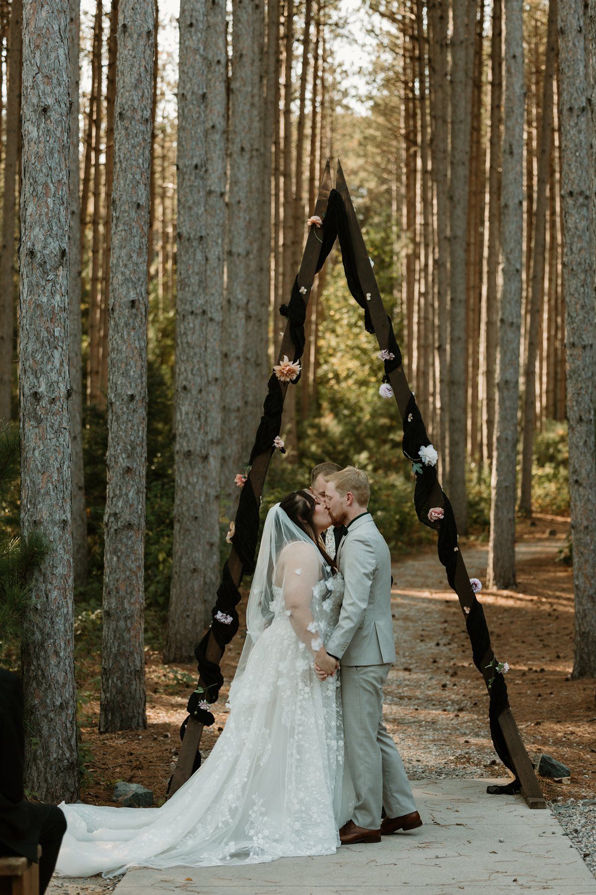 Bride and groom share first kiss under triangular wooden arch in pine forest wedding ceremony.