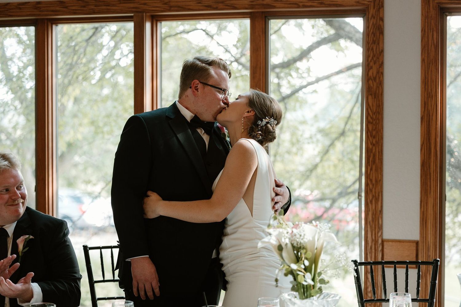 A couple shares a romantic kiss in a bright sunlit room with large windows.