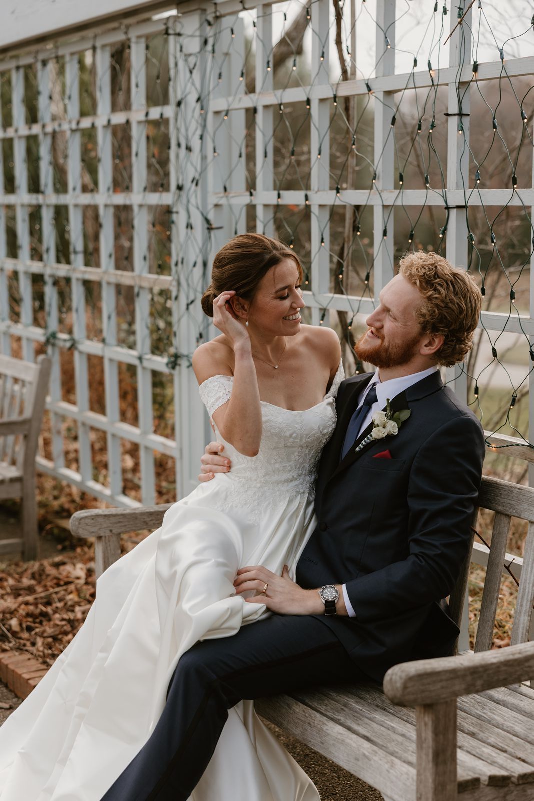 A romantic wedding couple shares an intimate moment while sitting on a wooden bench under a white lattice gazebo.