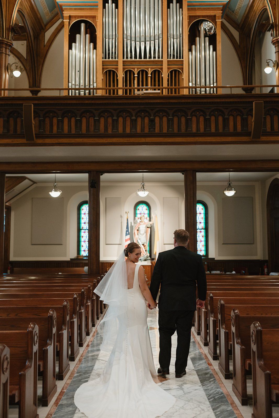A couple walks down the center aisle of a church with grand organ pipes visible above and stained glass windows.