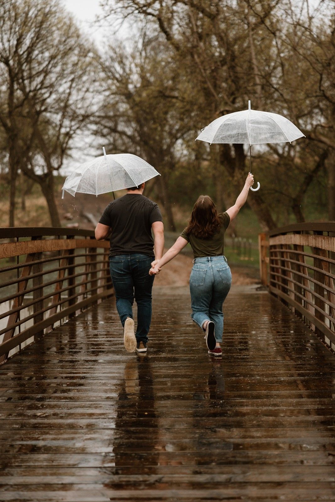 A couple walking across a wet wooden bridge holding clear umbrellas on a rainy day in nature.