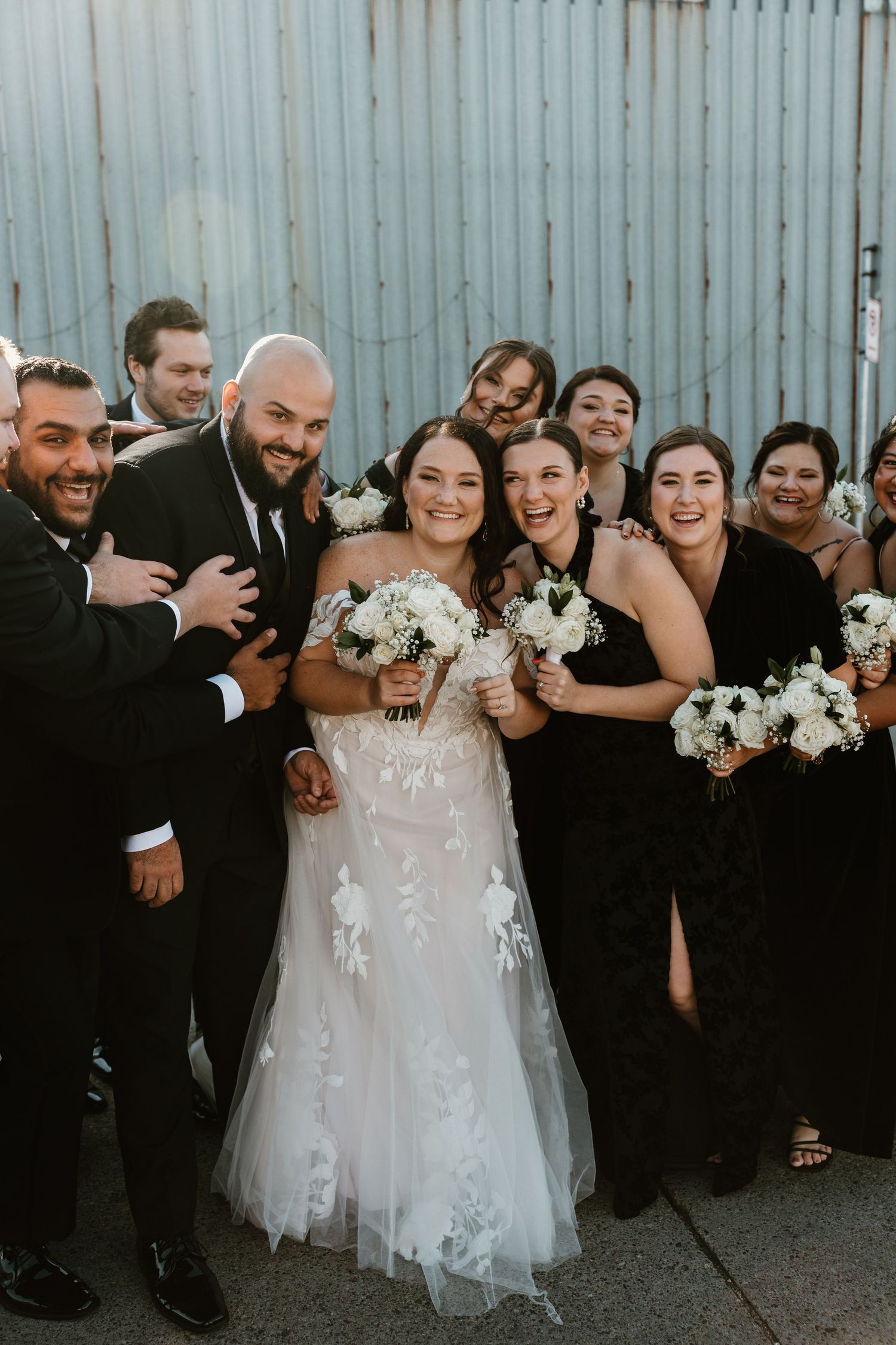 A wedding party in black attire stands together with the bride in a white floral lace gown holding white bouquets.