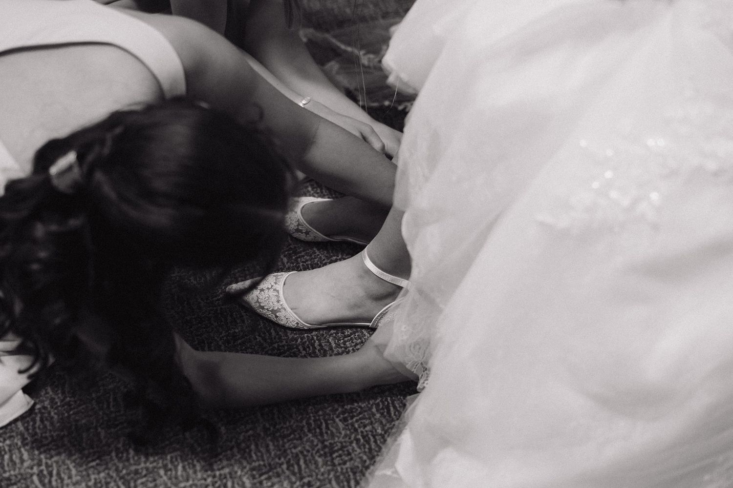 Black and white detail shot showing hands fitting a white ballet flat or bridal shoe on carpet.