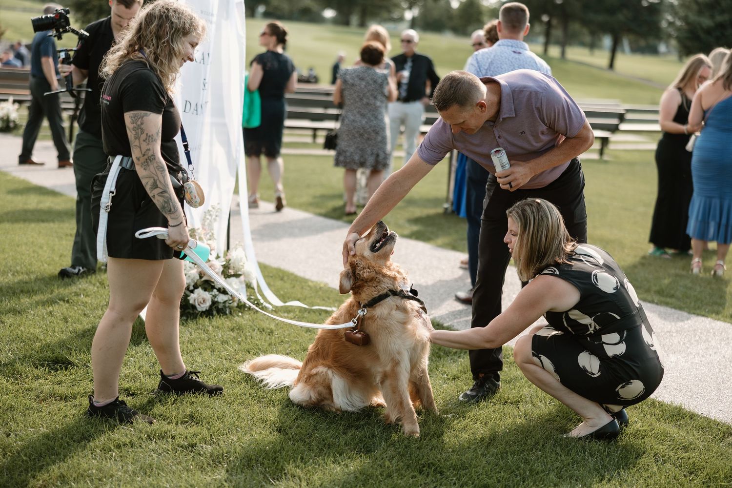 A golden retriever dog being petted by guests at an outdoor garden event on a sunny day.