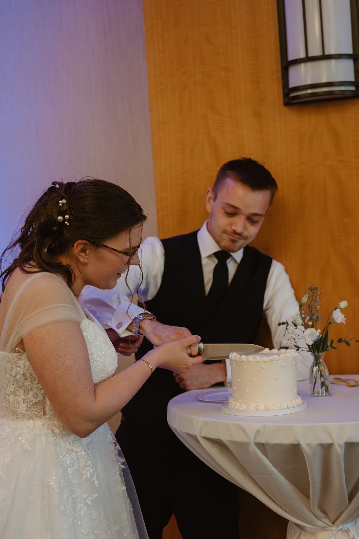 A newlywed couple cuts into their simple white wedding cake during their reception celebration.