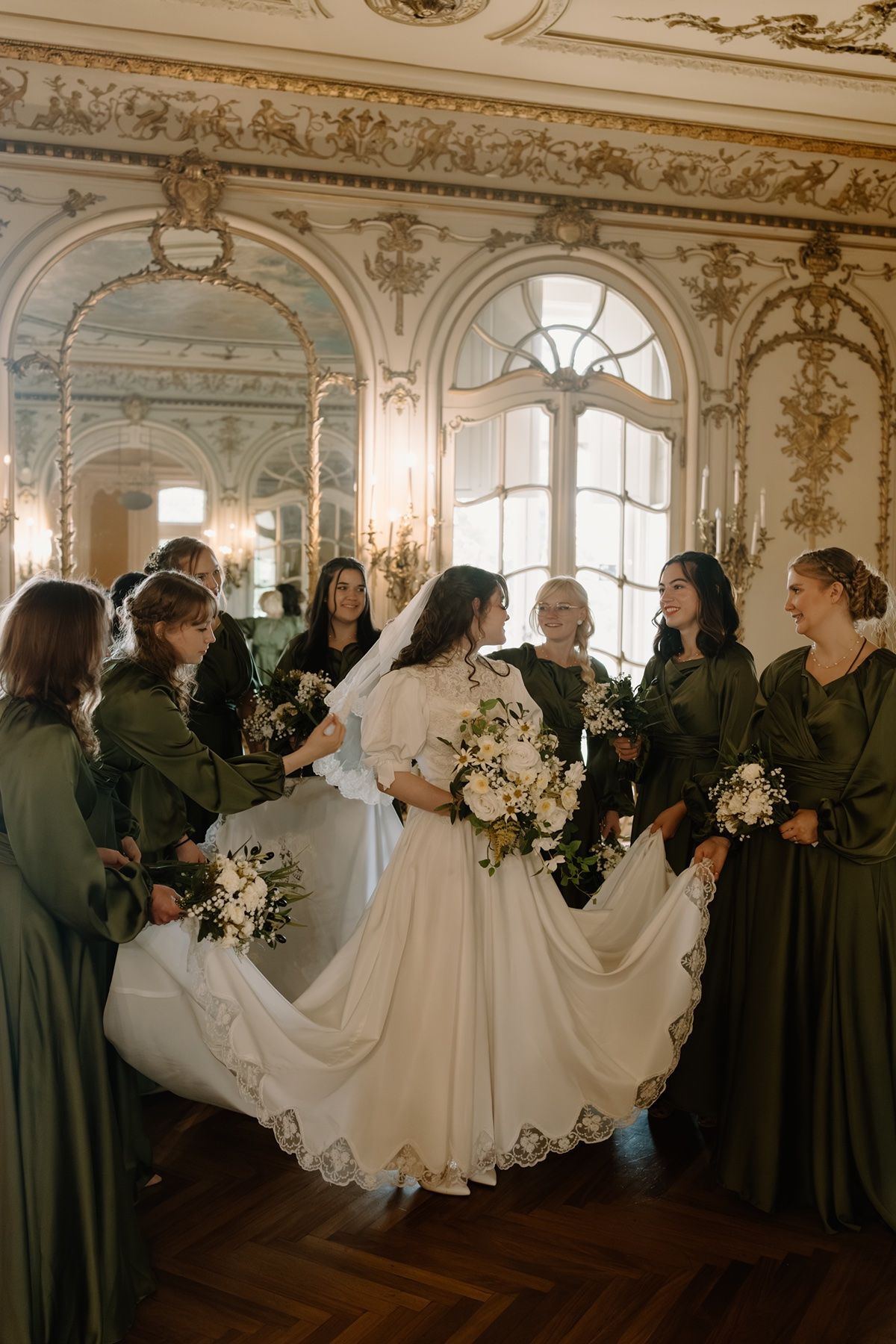 Bride in white gown twirls with bridesmaids in olive green dresses inside ornate baroque ballroom.