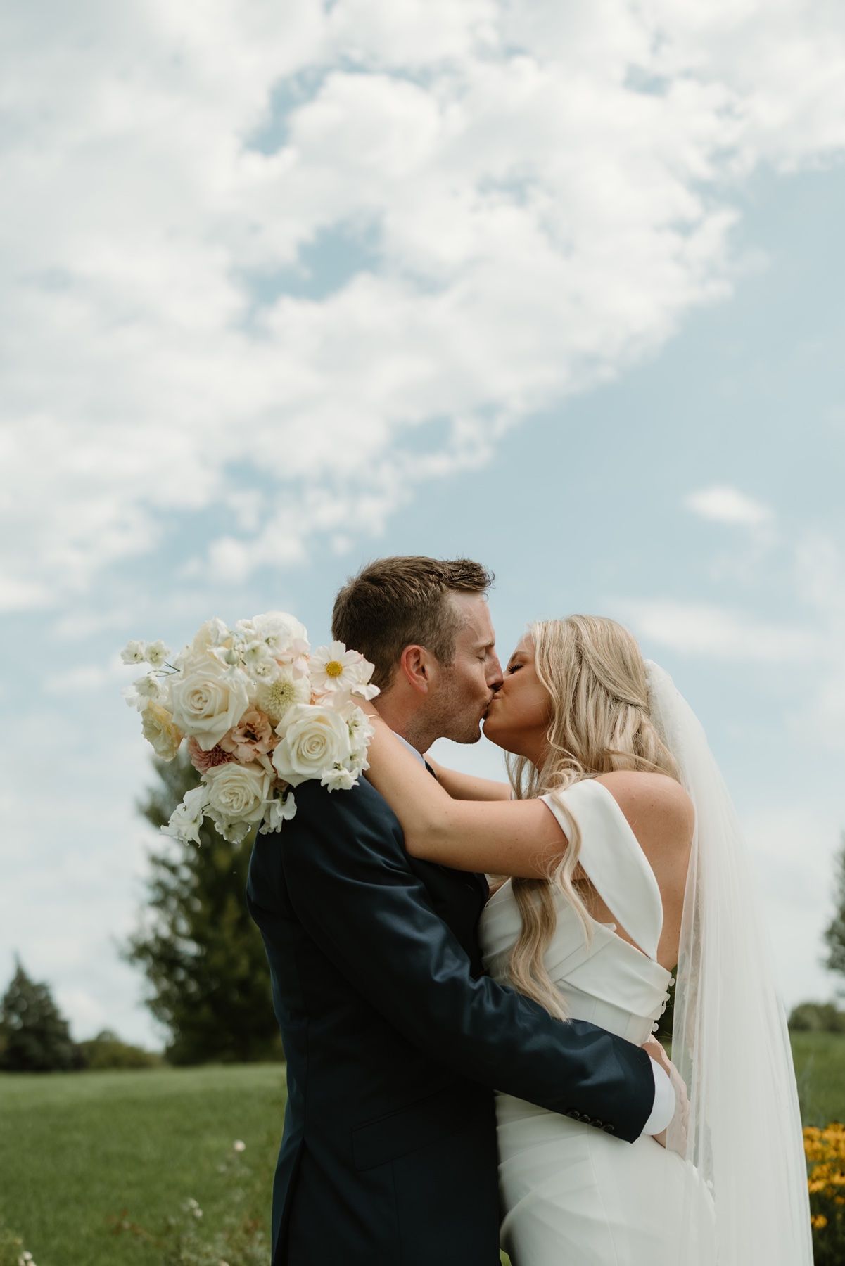 Bride and groom share a romantic kiss outdoors on their wedding day, with a white floral bouquet and cloudy blue sky backdrop.
