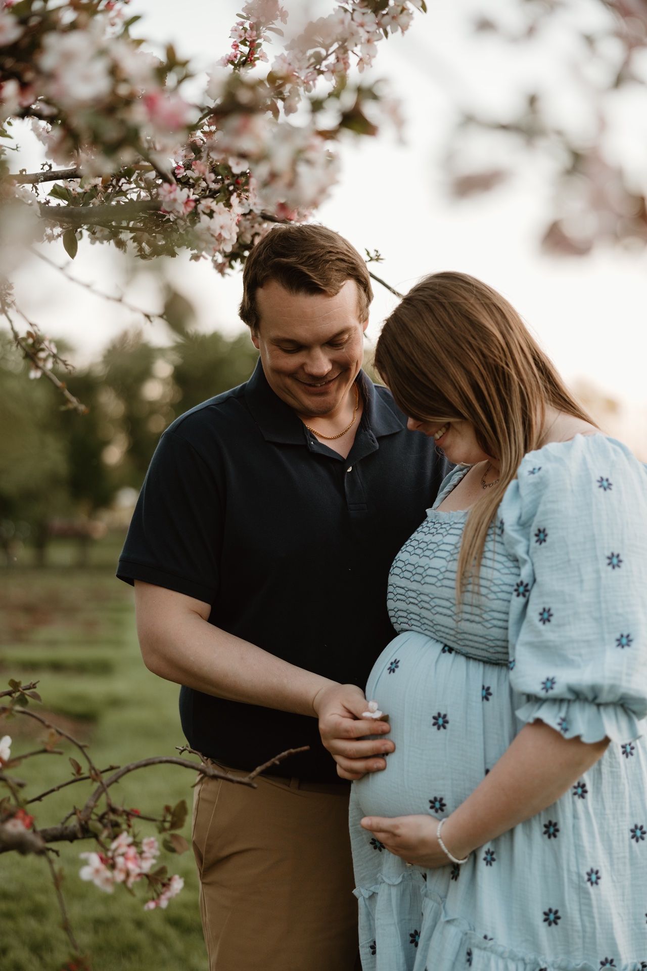 A smiling couple shares a tender moment outdoors under blooming pink flowers, the woman in a blue dress cradling her pregnant belly.