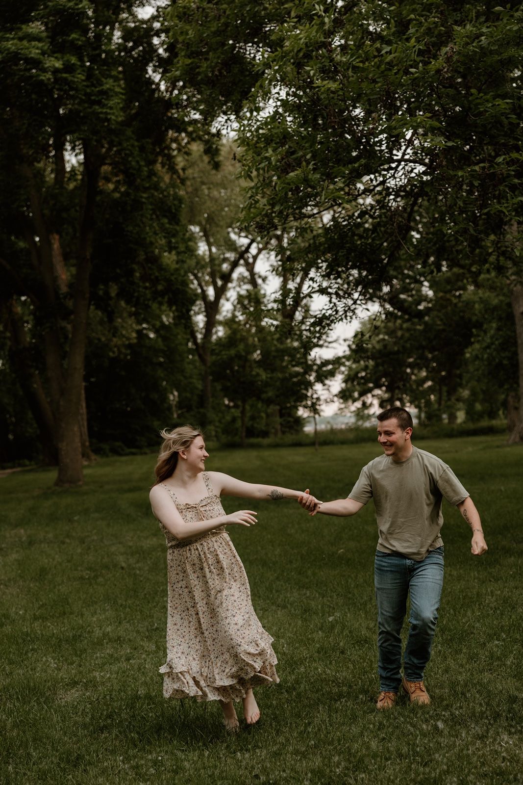 A couple holds hands and dances playfully in a lush green park, the woman wearing a floral dress.