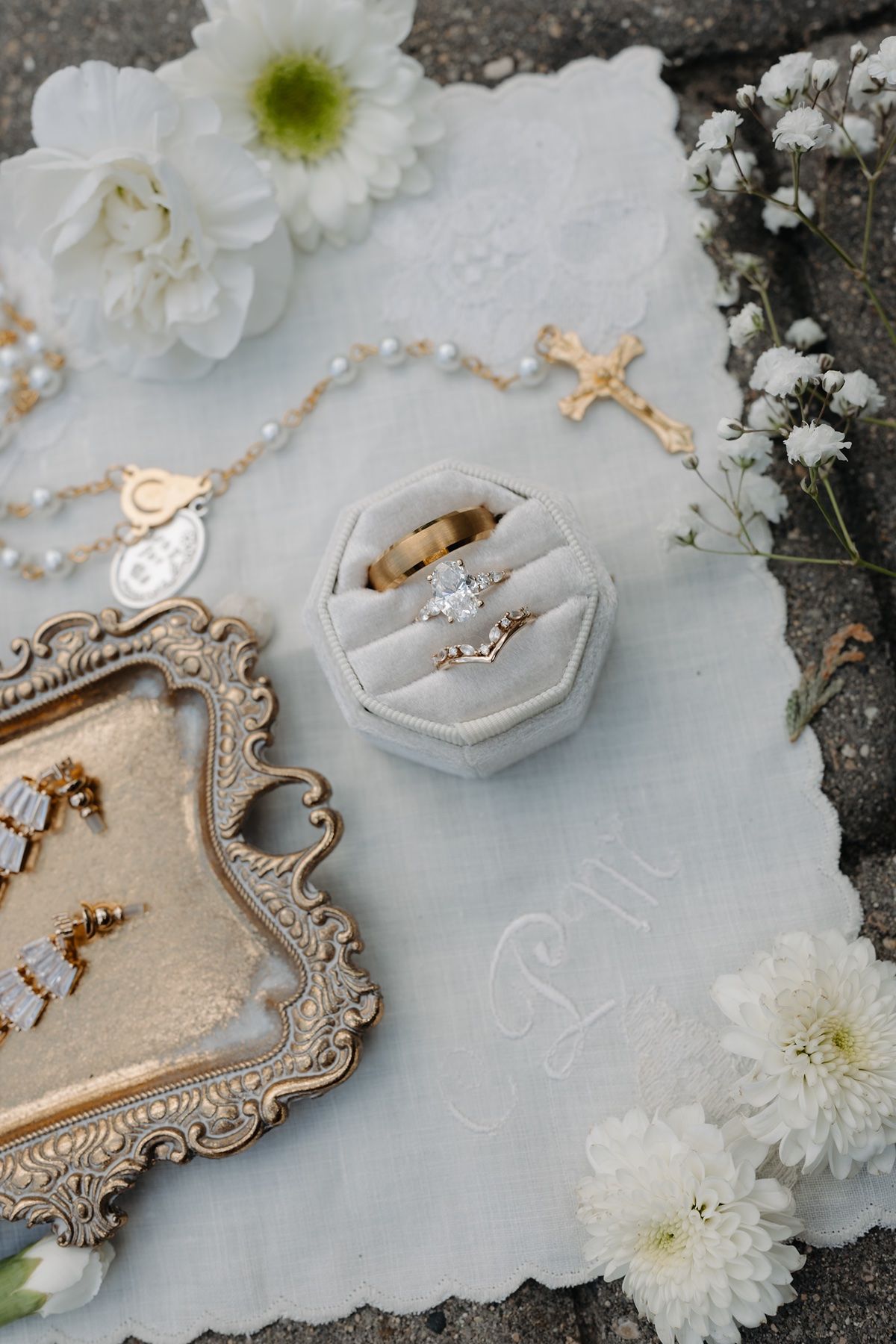 Elegant wedding flat lay with diamond engagement ring, gold band, rosary, earrings, and white flowers on linen.