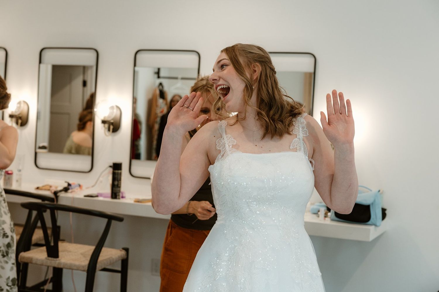 A person in a white wedding dress laughs joyfully while raising their hands in a bridal dressing room.