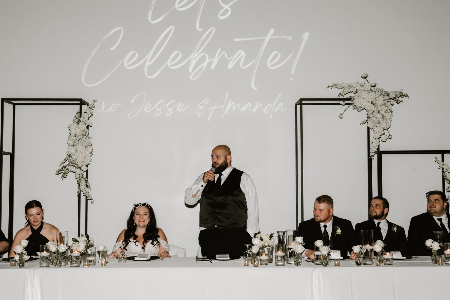 A person delivers a speech at a wedding reception head table decorated with white floral arrangements and formal place settings.