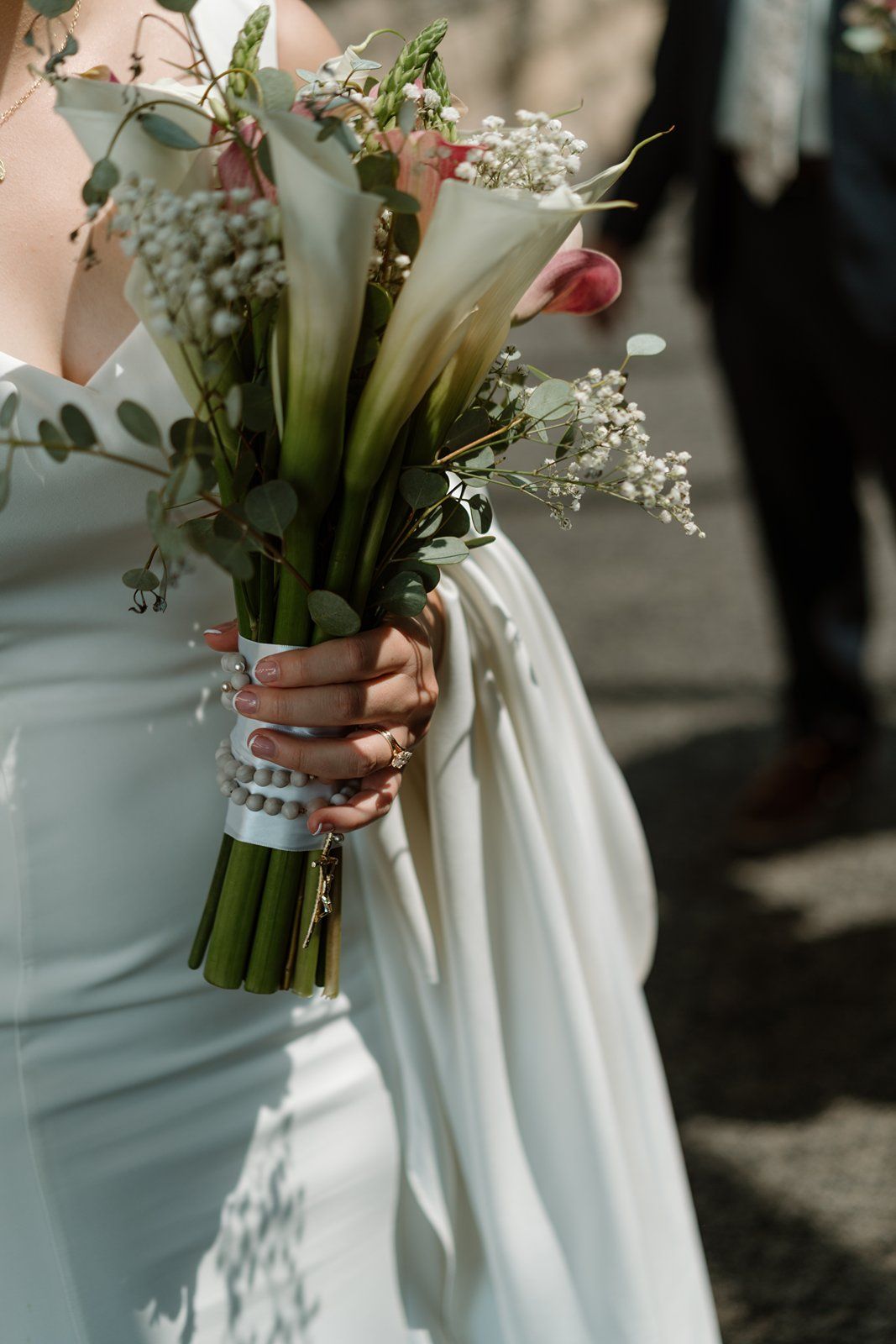 A delicate bridal bouquet of white calla lilies and baby's breath wrapped in satin ribbon held against a white dress.