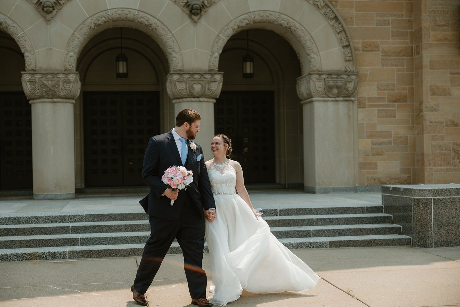 Newlyweds walk and pose together outside a grand stone building with arched columns and stairs.