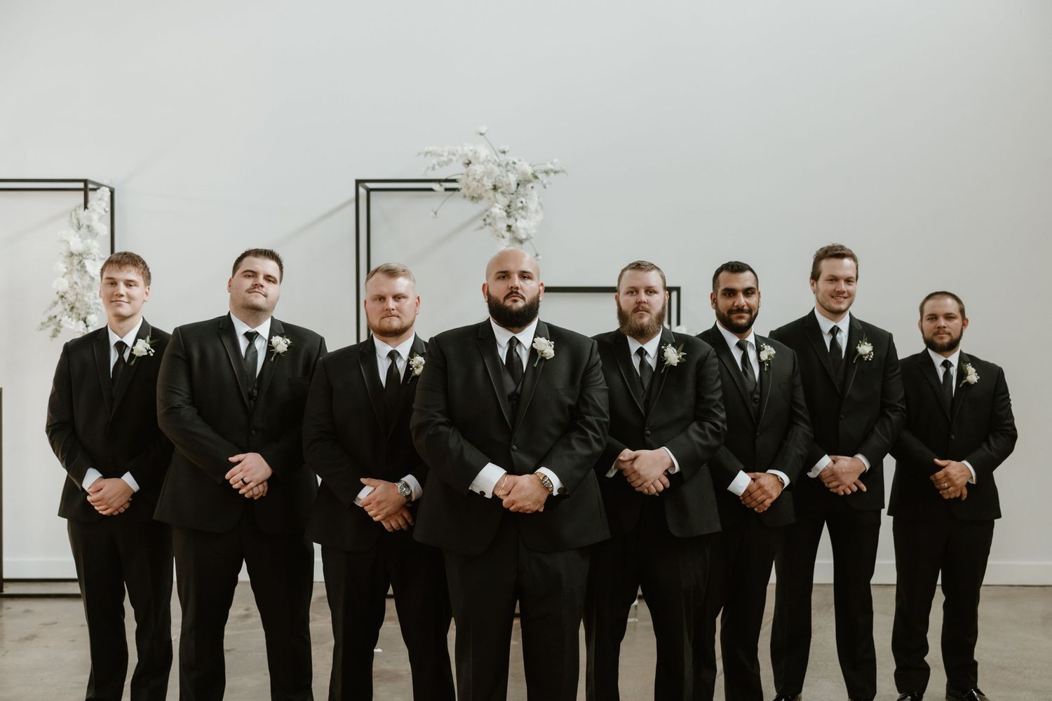 A row of groomsmen in matching black suits and ties stand together against a light colored wall at a wedding.