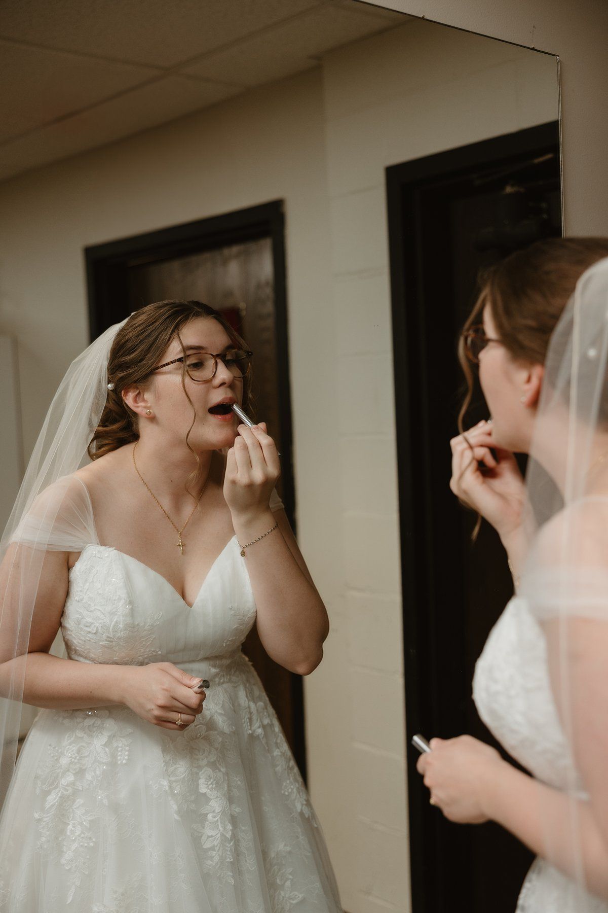 A bride wearing a lace wedding gown and glasses applies makeup while looking in a mirror.
