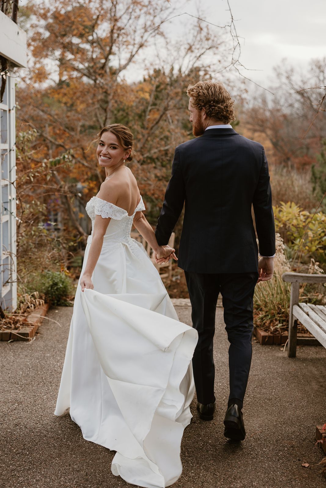 A bride in an off-shoulder gown walks with her groom along a garden path surrounded by fall foliage.
