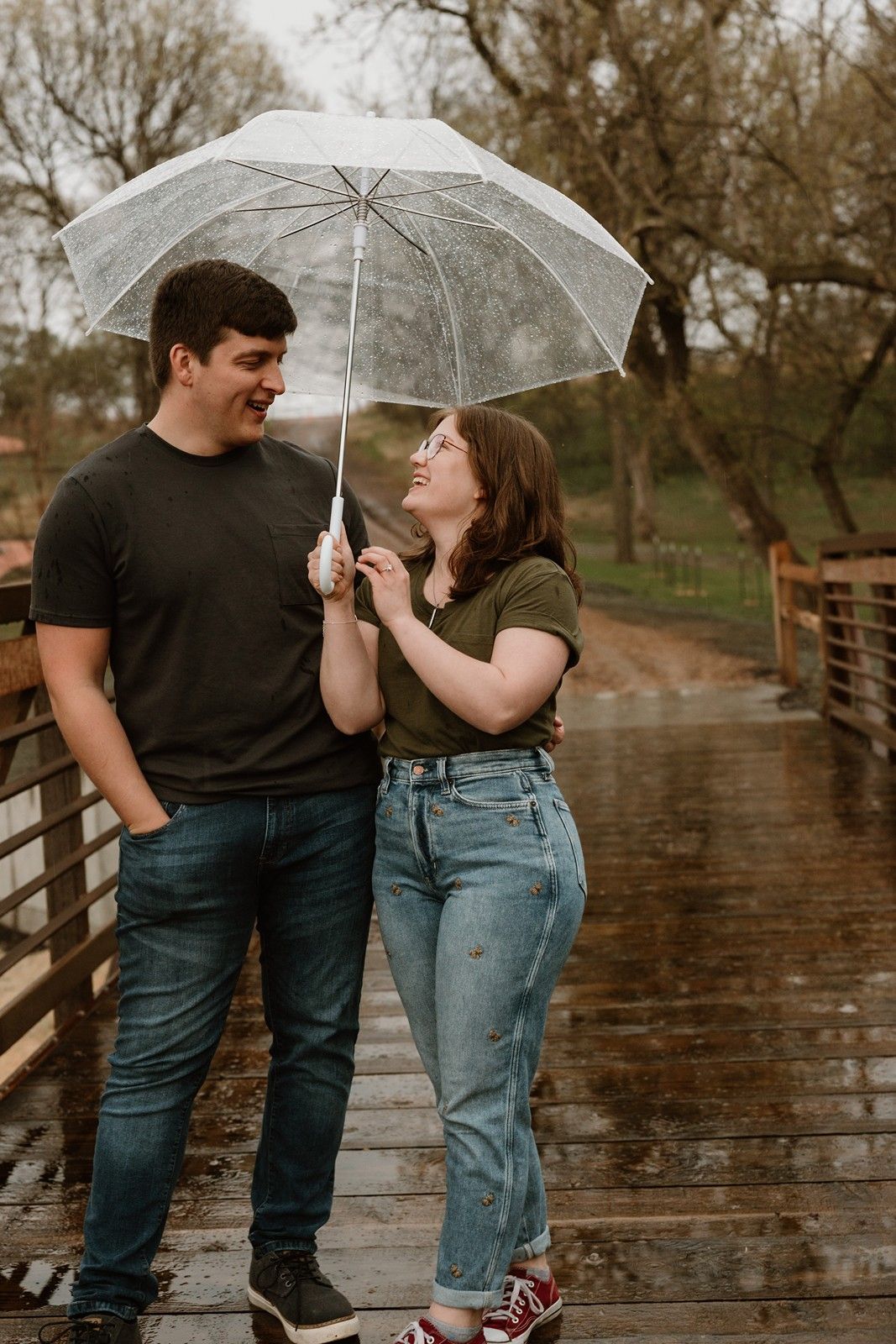 A couple shares a clear umbrella while standing on a wet wooden bridge in a rainy outdoor setting.