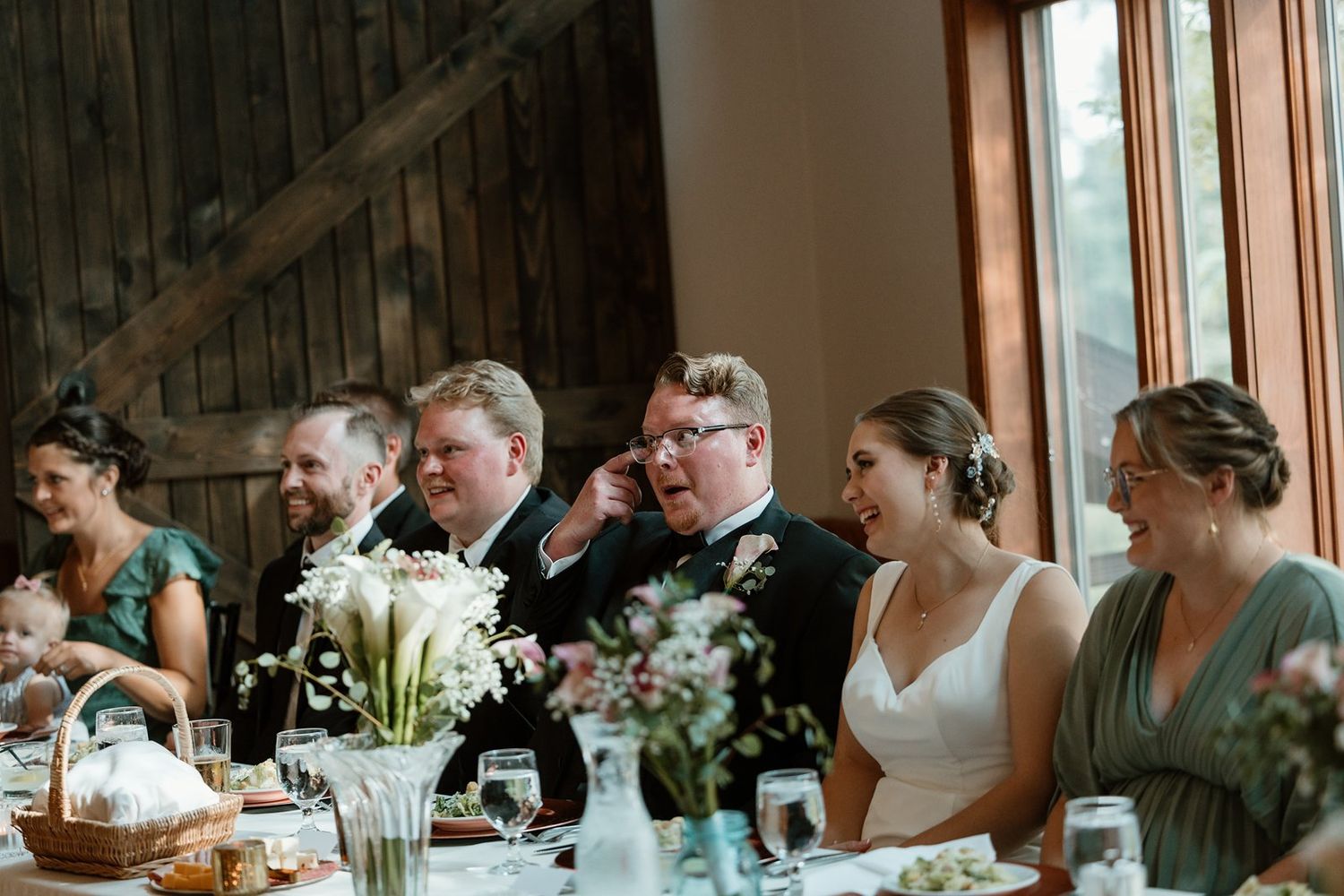 Wedding guests seated at long tables in a rustic barn venue with natural lighting and floral centerpieces.