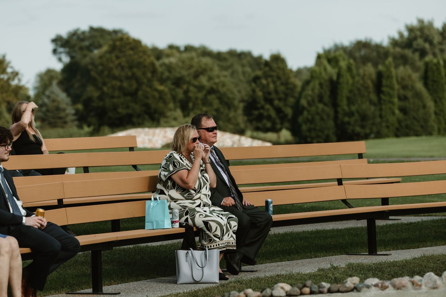 Wedding guests sit on wooden benches arranged outdoors for a ceremony in a natural setting.