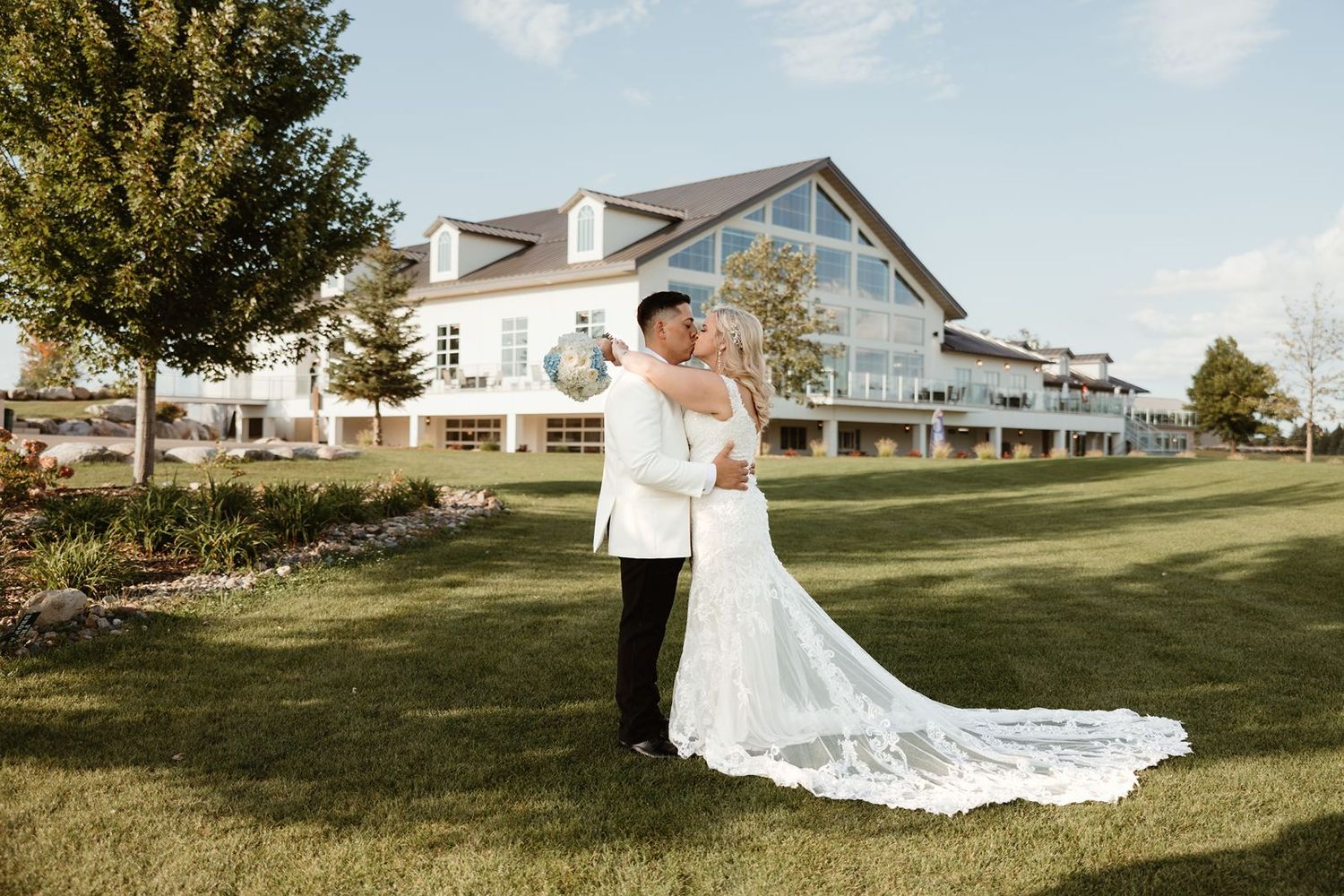 A wedding couple poses in front of a grand white venue on a sunny day.