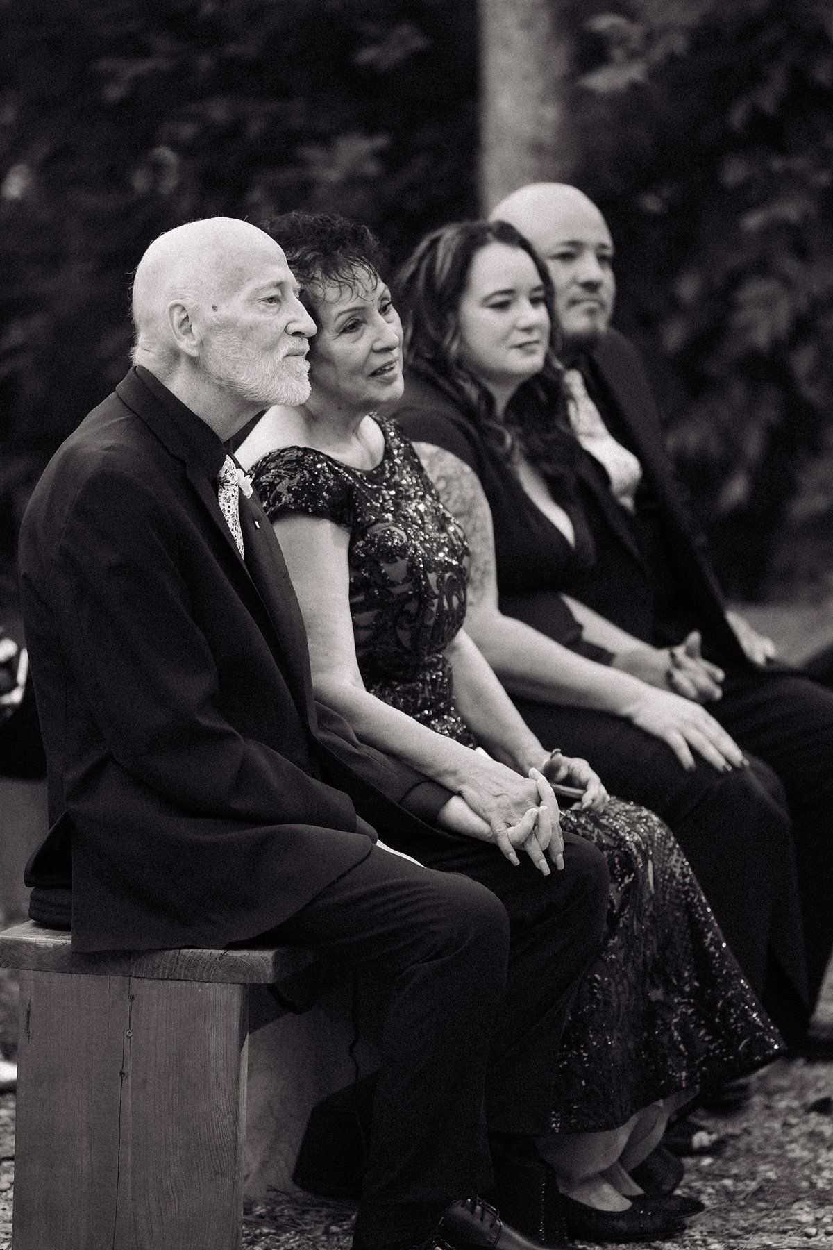 Black and white photo of four formally dressed guests seated on a bench at an outdoor wedding ceremony.