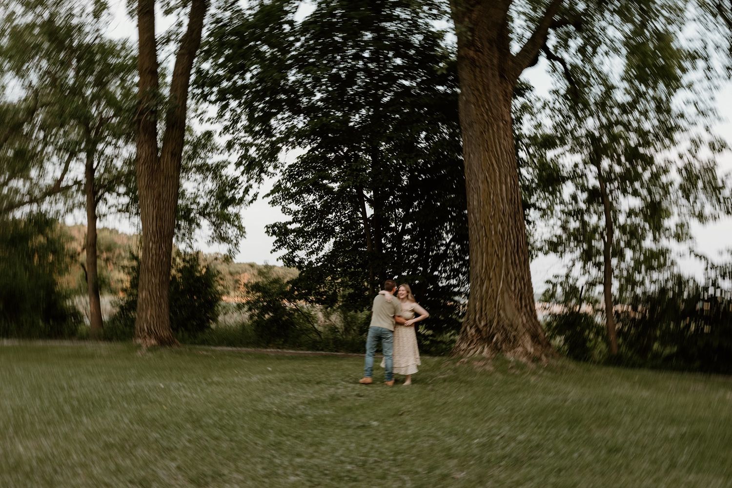 Couple embracing between tall trees in a lush green park at dusk during a romantic outdoor engagement session.