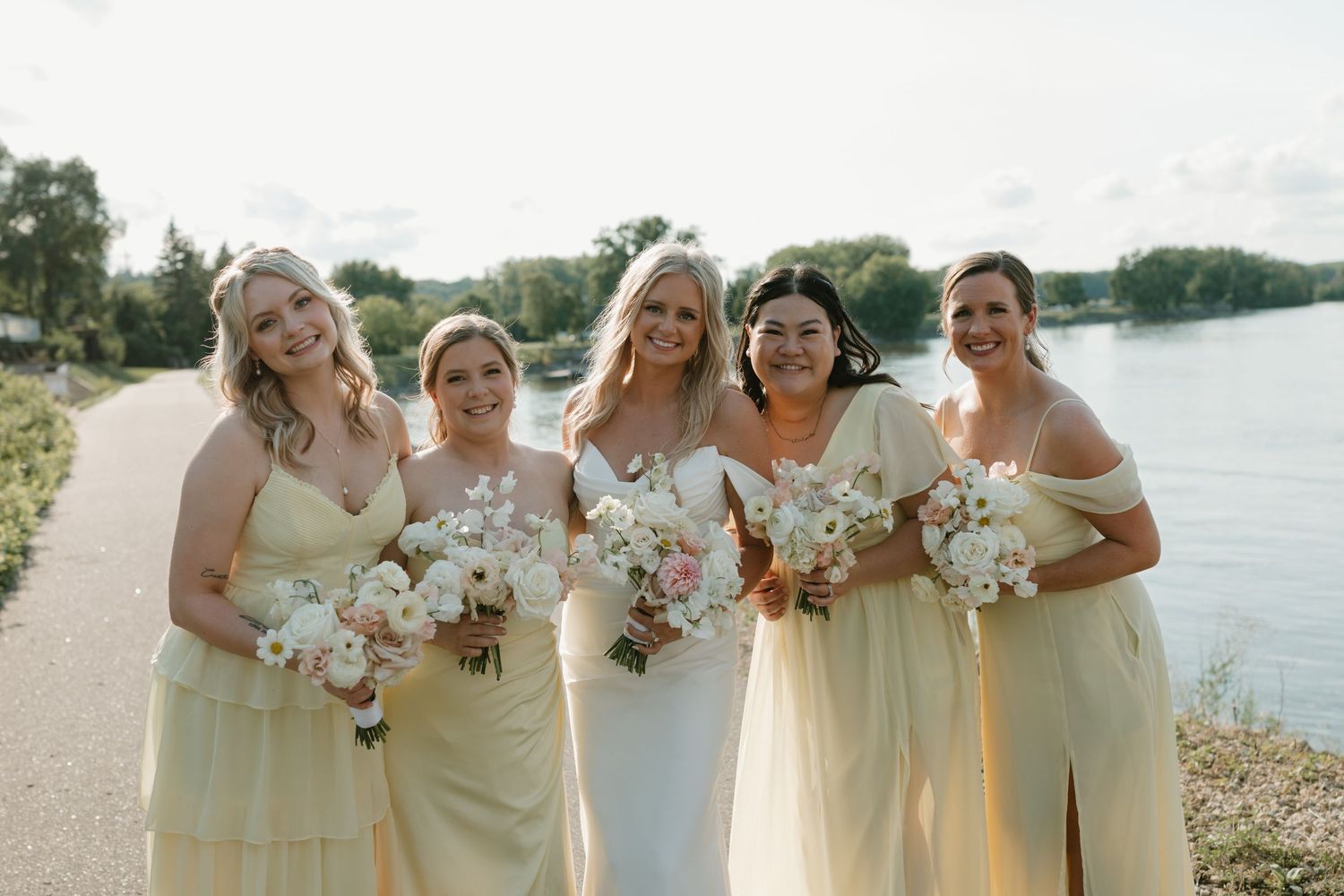 Wedding party in pale yellow bridesmaid dresses holding white bouquets pose by waterfront on sunny day.