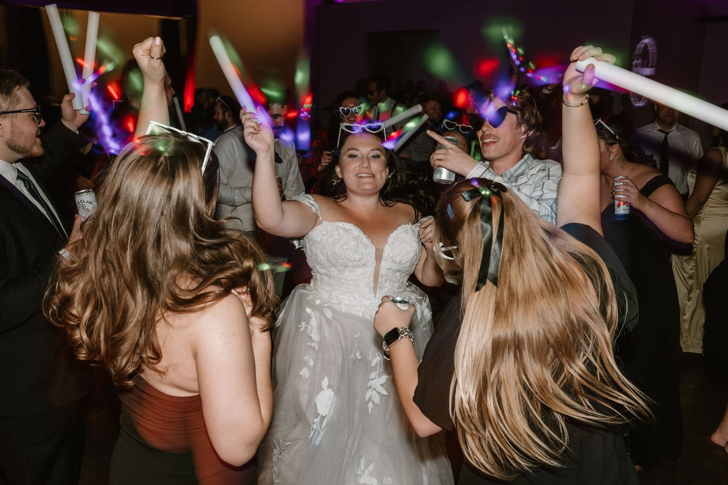 Wedding reception party guests dance with glowing LED light sticks on a dark dance floor.