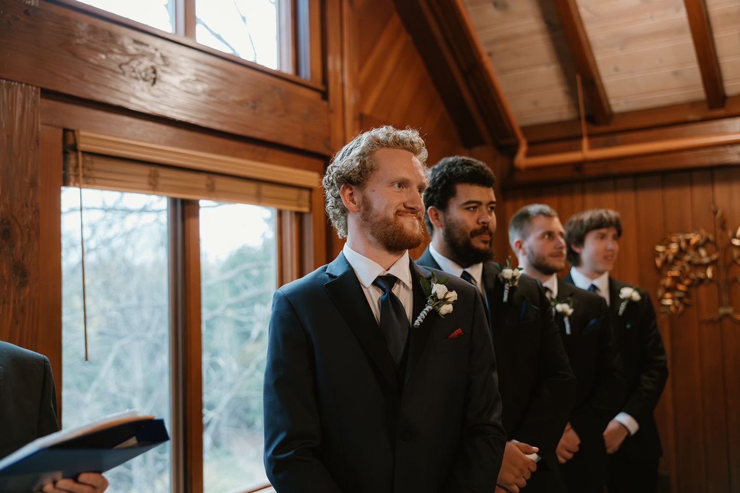 Groomsmen in dark suits stand at attention during a rustic wooden chapel wedding ceremony.