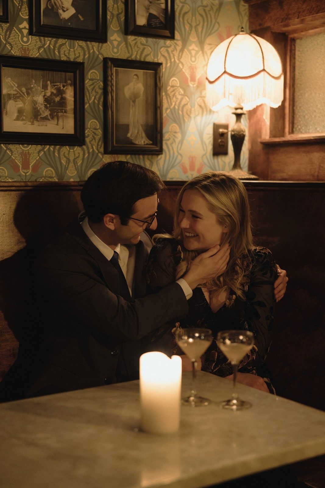 A romantic candlelit moment between a couple sharing wine in a vintage-style restaurant with ornate wallpaper and artwork.