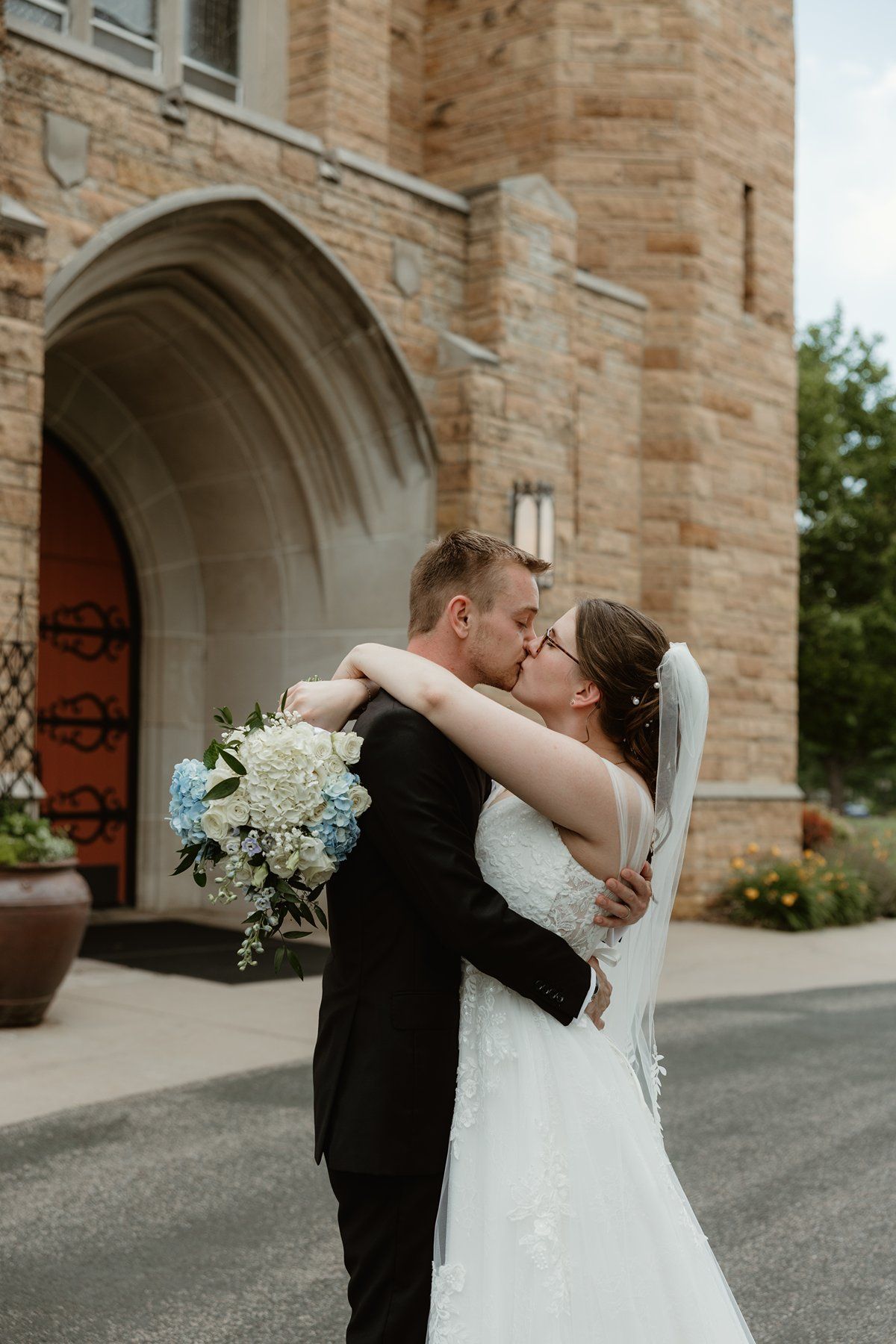 A romantic wedding couple shares kisses in front of a stone church archway with white floral bouquet.
