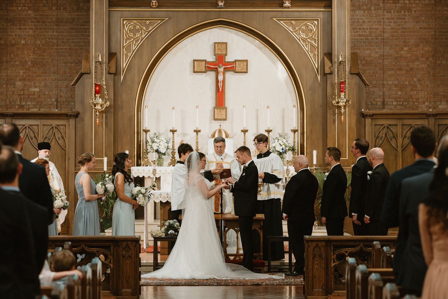 Elegant church wedding ceremony with bride and groom at altar beneath ornate crucifix, flanked by wedding party.