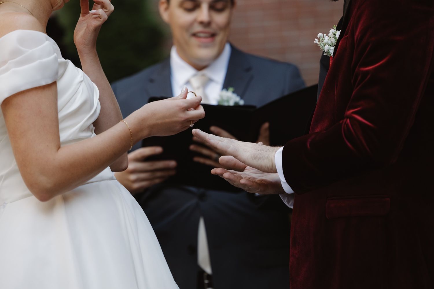 Bride placing wedding ring on groom's finger during outdoor ceremony with officiant in background.