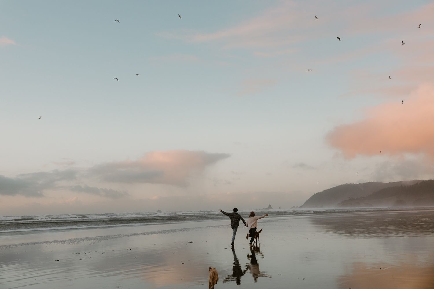 Couple joyfully running on a reflective beach at sunset with birds flying overhead and misty coastal hills in the background.