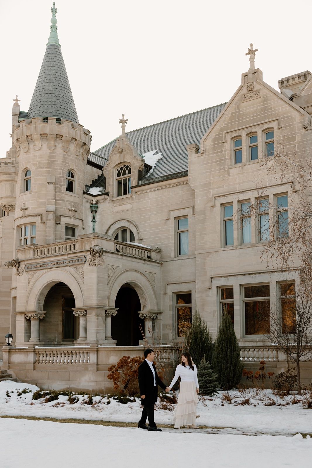 Couple holding hands in snow outside a grand castle-like stone mansion with turret in winter.