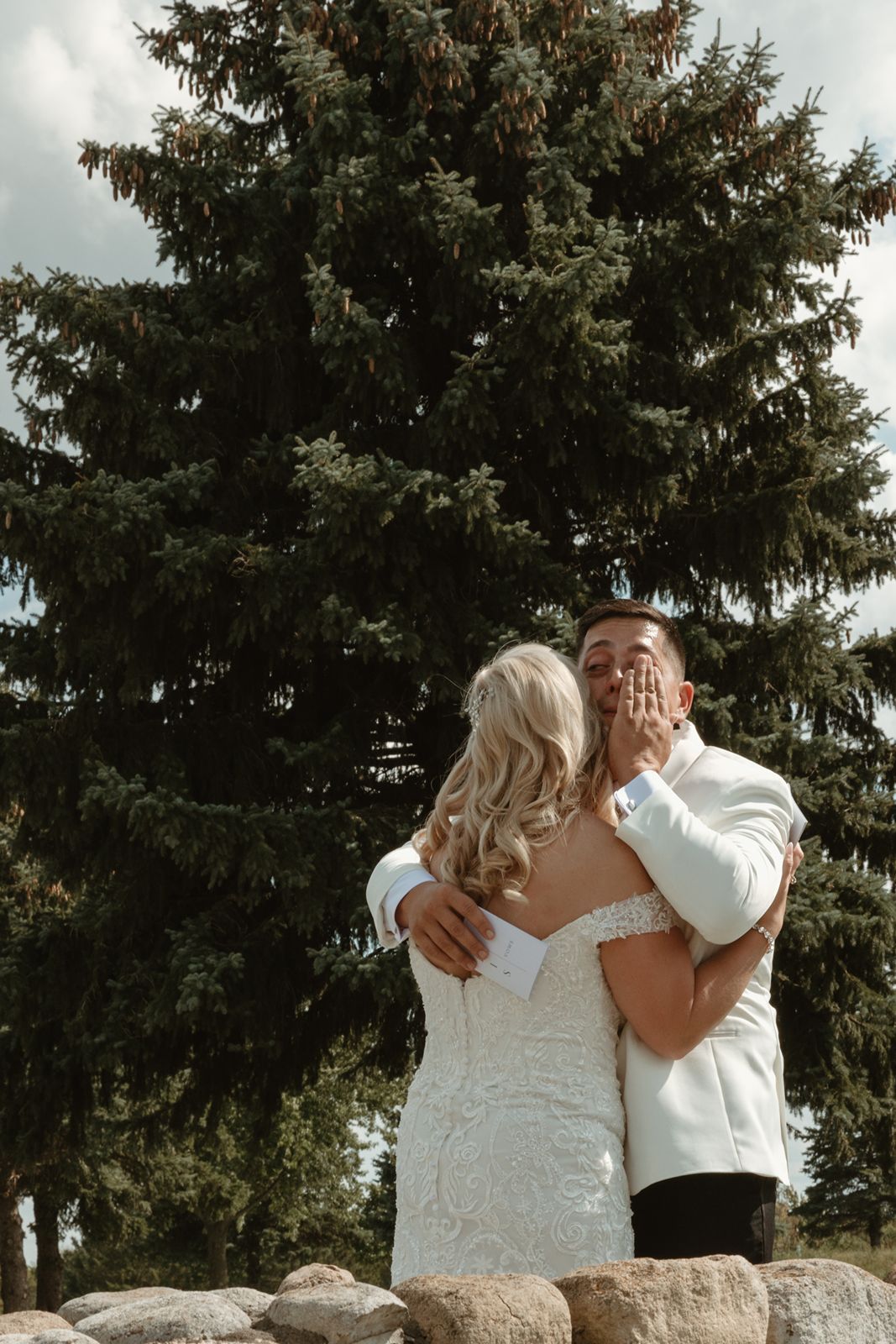A tender wedding moment captured in front of a towering evergreen tree.