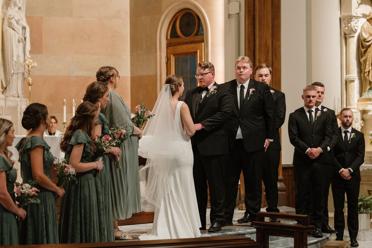 Wedding ceremony taking place in a church with bridesmaids in sage green dresses and groomsmen in black suits.